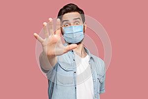 Stop, don`t come any closer. . Portrait of angry young man with surgical medical mask in blue shirt standing with stop hand sign