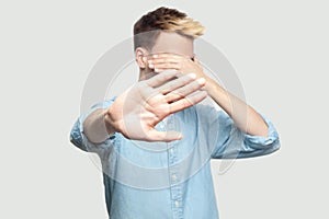 Stop, I don`t want to see at this. Portrait of scared handsome young man in light blue shirt standing, covering his face and