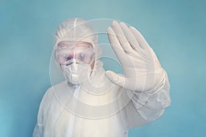 Stop the infection. Doctor with full body suit and mask showing stop sign with his hand on blue background. Corona virus or covid-