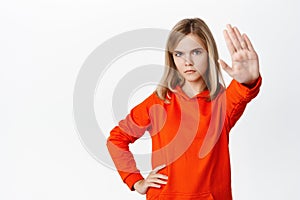 Stop. Little girl looks serious, extends one arm to say no, rejecting, prohibit action, forbid smth, standing in red photo