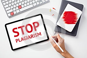 Stop plagiarism concept. Office desk table with keyboard, paper notebooks, tablet. Message STOP PLAGIARISM on tablet screen