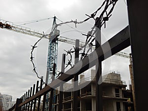 Stopped high-rise construction crane behind a metal fence with barbed wire in focus on foreground