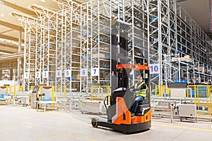 Storehouse employee in uniform working on forklift in modern automatic warehouse. Boxes are on the shelves of the warehouse.