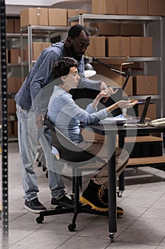 Storehouse workers looking at delivery logistics on laptop computer analyzing checklist searching for metallic box