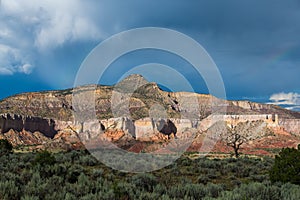 Storm clouds and faint rainbows over a desert mountain peak with colorful cliffs highlighted by the light of sunset