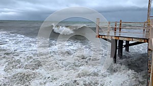 Storm on the sea. Waves cover the pier. Thunderclouds in the sky. Strong wind on the ocean. Autumn on vacation. Old