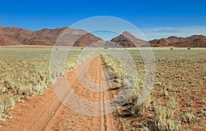 Straight desert dirt road track passes grassland towards mountains.