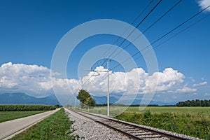 Straight railroad tracks between agriculture fields with parallel countryside asphalt road in Slovenia