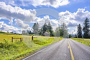 Straight road along a summer meadow with lush grass and an old rickety wooden barn at the edge