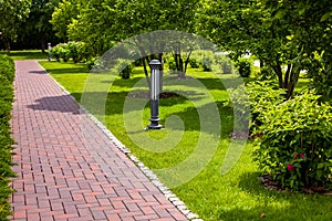 Straight stone tile pavement in a park landscaped with green grass and bushes.