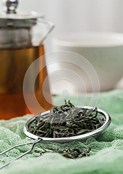 Strainer infuser with loose green organic tea on green cloth with clear glass teapot