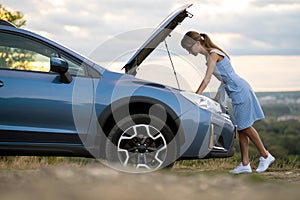 Stranded young woman driver standing near a broken car with popped up bonnet inspecting her vehicle motor