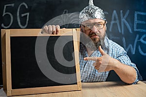 A strange bearded conspiracy theorist in a protective aluminum foil hat and glasses sits at a table and points his