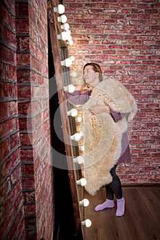 Strange ugly aged woman in in a fur Cape in front of a lighted mirror in a dark room. Old model posing for a photo shoot in the
