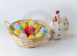 A straw basket, colored Easter eggs lying in it, a burning red candle, and a ceramic figure of a chicken