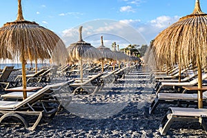 Straw beach umbrella with blue sky, sunbeds on the black volcanic beach of La Caletta. Tenerife, Canary Islands, Spain