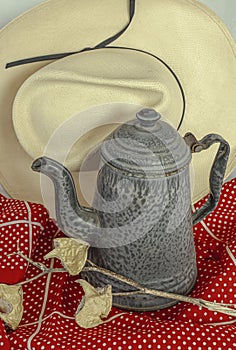 Straw cowboy hat frames metal granite ware tea coffee pot. Red polka dot tablecloth adds color.