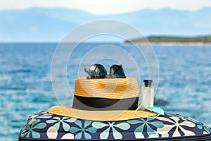 A straw hat on a bag with a mask, hand sanitiser and sunglasses over beach background.