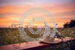 A straw hat and a bouquet of wild flowers on a wooden bench in a field at sunset