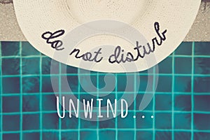 Straw hat with Do not disturb wording at the pool edge with Unwind... wording