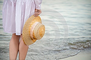Straw hat in female hand on seashore at sunset. Unrecognizable Woman in white beach dress having rest at sunset on sea beach.