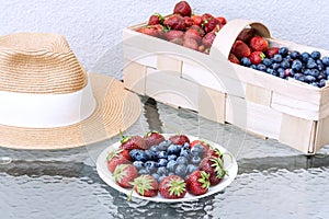 A straw hat, fresh strawberries and blueberries lie in a plate and basket on a glass table against the background of a plastered