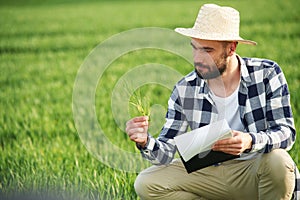 In straw hat. Holding wheat and notepad. Handsome young man is on agricultural field