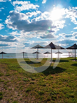 Straw umbrellas on green grass harsh shadow, sand beach vivid blue sea water sky background sunbeam. Scenic landscape