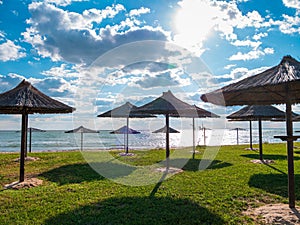 Straw umbrellas on green grass harsh shadow, sand beach vivid blue sea water sky background sunbeam. Scenic landscape