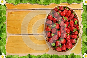 Strawberries in basket on wooden table with a frame of strawberry leaves and flowers - birds eye view