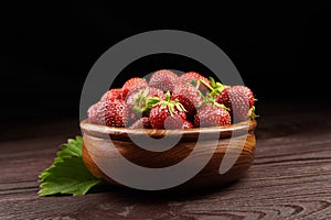 Strawberries with green leaves in wooden bowl on wooden table. Red ripe berries, fresh juicy strawberries on dark background