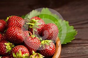 Strawberries with green leaves in wooden bowl on brown wooden table. Red ripe berries