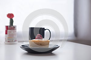 A strawberry cheese tart , a black cup of hot coffee and cactus on white vintage wooden table and curtain background