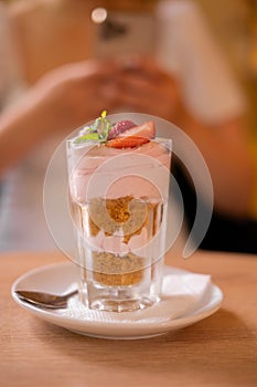 Strawberry dessert tiramisu in glass on cafe table and woman with phone on background, selective focus, summer