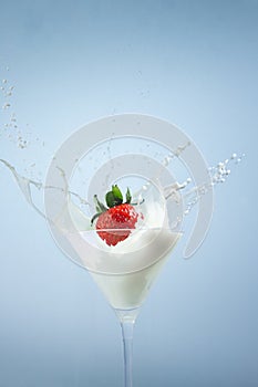 Strawberry dropping into a cocktail glass of milk creating splashes on blue background