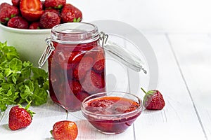 Strawberry jam in a glass jar next to fresh strawberries. On a white wooden background. Homemade winter fruit blanks. Selective
