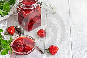 Strawberry jam in a glass jar next to fresh strawberries. On a white wooden background. Homemade winter fruit blanks. Selective