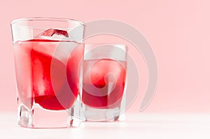 Strawberry red cocktails in two shot glasses with ice cubes closeup on white wood table and pastel pink wall, copy space.