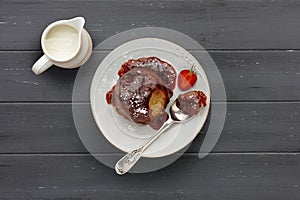 Strawberry sponge cake, with a sticky strawberry sauce, on a white plate with a spoon and a jug of cream, on a dark grey wooden
