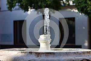 Stream of crystal clear water gushing out of stone fountain. In the background white walled house in mediterranean style in spain