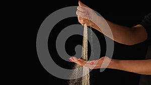 Stream of dry sand pours into palm of woman and is spilling through her fingers on black background. Small grains of