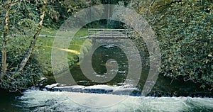 Stream with rapid current near a bridge, surrounded by trees. Kilbrittain Woods. pano