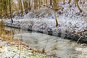 Stream with sun reflection on water surface next to snowy hill with wild vegetation