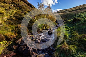 Stream with tree in the center, meadows and hills on sides, beautiful autumn colors with blue sky and puffy clouds