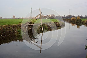 Stream of water in the countryside and a mooring pole by the shore with its reflection casted in the water on a cloudy day