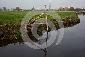 Stream of water in the countryside and a mooring pole by the shore with its reflection casted in the water on a cloudy day