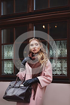 Street fashion concept: portrait of young beautiful woman wearing pink coat with handbag posing at the window. City