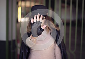 Street fashion outdoor photo of woman with dark hair in black leather jacket and sunglasses in defocus. Woman gesturing STOP with