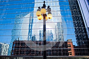 Street lightpost shines in front of a reflective office building, which mirrors the images of surrounding skyscrapers