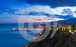 Street lights of Giardini Naxos near Taormina reflected in serene Ionian sea. Sunset view of coast of Sicily, Italy